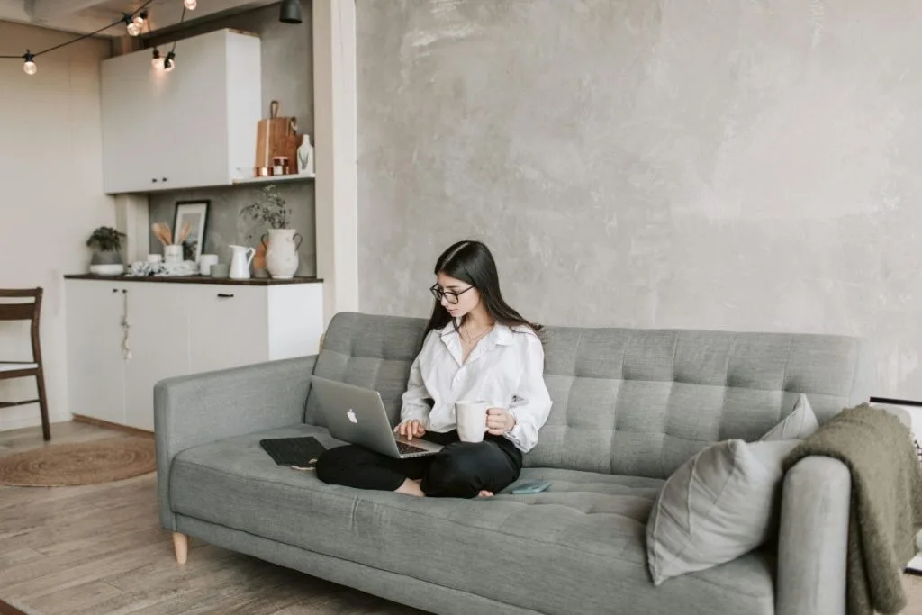 Chica intentando recuperar el dinero que ha sido estafada por Internet, sentada en un sofá con el portátil y una taza en la mano.